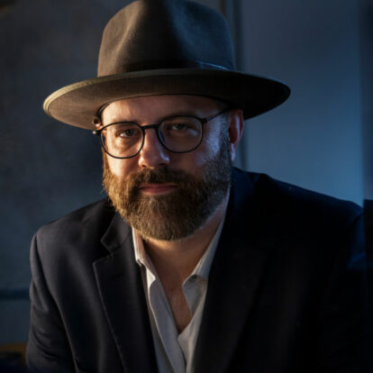 Musician Afton Wolfe in a wide-brimmed hat and glasses, wearing a dark blazer in a moody, dimly lit setting.