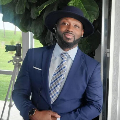 DJ Desmond Newsom in a navy-blue suit and patterned tie, confidently posing against a lush green backdrop.