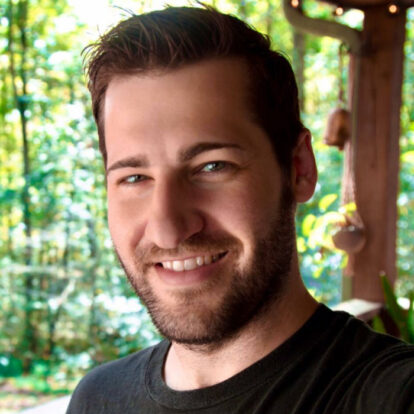 DJ Tanner Roman in a dark flannel shirt and cap, grinning while posing indoors.