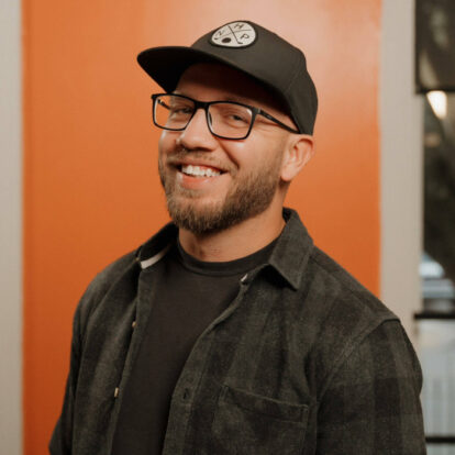 Comedian Matt Taylor with a beard, black cap, and glasses, smiling warmly in front of an orange wall.